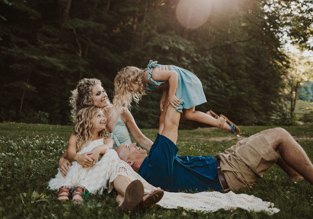 Family sitting on ground together.  Daughter leaning into mother's arms while dad holds other daughter up in the air